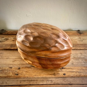 May include: A hand-carved wooden box shaped like a large walnut, featuring a rich brown finish with a textured, dimpled surface. The object consists of two halves that fit together, showcasing the natural wood grain and a polished appearance.