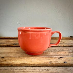 Puede incluir: Una taza de cerámica de color coral sólido con forma de cuenco ancho y redondeado, un asa lateral resistente y dos anillos horizontales en relieve que rodean la parte superior del exterior. La taza tiene un acabado liso y brillante.