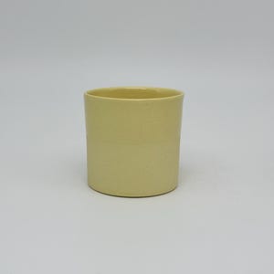Könnte beinhalten: Eine zylindrische, hellgelbe Keramiktasse. Die Tasse hat eine glatte, matte Oberfläche und ein schlichtes, minimalistisches Design. Die Tasse ist etwa 10 cm hoch und 7,6 cm im Durchmesser, geeignet für Getränke oder als Dekorationsartikel.