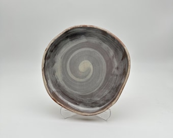 May include: A handmade ceramic plate with a unique, organic shape. The plate features a spiral design in shades of gray and brown, with a light beige center. The rim has a darker brown color. The plate is displayed on a clear stand.