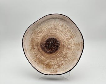 May include: A decorative, organic-shaped bowl with a light brown interior and a dark brown center. The bowl has a black rim and sits on a clear stand. The interior features a textured, concentric circle design.