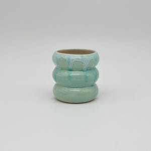 May include: A small, handmade ceramic vase in a stacked, rounded design. The vase is a soft, seafoam green color with a light blue glaze dripping down the top. The interior is a neutral tan color. The vase is suitable for small plants or as a decorative item.