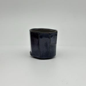 Puede incluir: Una taza de cerámica azul oscuro con un diseño geométrico único. La taza tiene un exterior facetado y un borde ligeramente irregular. El esmalte tiene una apariencia sutil y texturizada. La taza está diseñada para bebidas.