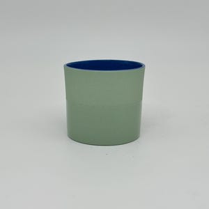 May include: A cylindrical cup with a sage green exterior and a deep blue interior. The cup has a simple, minimalist design, with a clean, smooth surface. The interior color provides a striking contrast to the exterior. The cup is likely made of ceramic or a similar material.