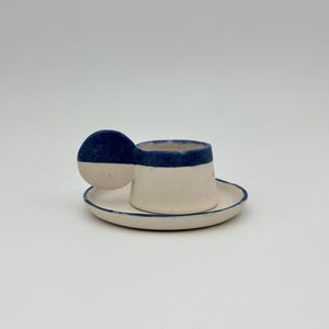 Könnte beinhalten: Ein kleines, handgefertigtes Keramik-Tassen- und Untertassen-Set. Die Tasse ist cremefarben mit einem dunkelblauen Rand und Henkel. Die Untertasse hat einen passenden blauen Rand. Das Set ist für Espresso oder ein kleines Getränk konzipiert.
