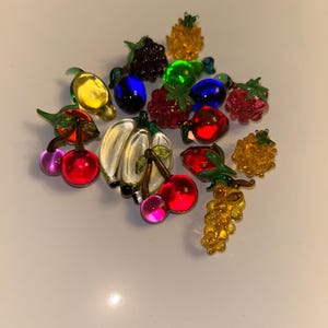 May include: A collection of colorful glass fruit ornaments, including cherries, grapes, bananas, and berries. The ornaments are in various colors, such as red, yellow, green, and purple. The fruit is arranged on a white surface.