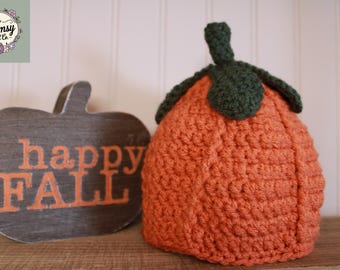 Pode incluir: Uma abóbora laranja de crochê com caule e folhas verdes está ao lado de uma placa de madeira em forma de abóbora com a inscrição "happy FALL" em letras laranja. Decoração de outono.