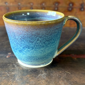 May include: A handmade ceramic mug with a wide, slightly flared shape. The mug features a gradient of colors, transitioning from a golden-brown rim to a deep blue body. The handle is a matching golden-brown color.