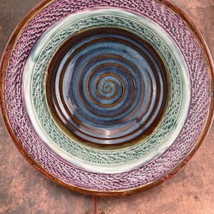May include: A large, round ceramic bowl with a spiral design in shades of blue, brown, and green. The bowl has a textured rim in a purple hue.