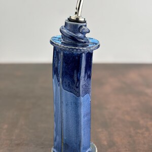 May include: A handcrafted, blue ceramic oil bottle with a stainless steel spout. The bottle has a unique, octagonal shape and a textured, glazed finish. The top features a decorative swirl design. The bottle measures approximately 8 inches tall.