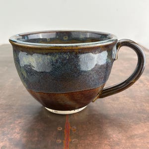May include: A handmade ceramic mug with a blue, brown, and white glaze. The mug has a wide, rounded body and a handle.