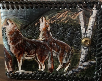 May include: A black leather wallet with a tooled design of two wolves howling at the moon in a forest setting. The wallet has a snap closure.