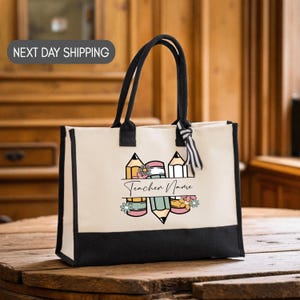 May include: Cream-colored tote bag with black trim and handles. The bag features a colorful pencil and flower design with the text "Teacher Name". The bag is sitting on a wooden surface.