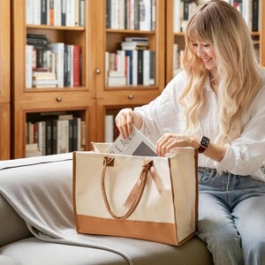 A woman with long blonde hair is sitting on a couch, holding a book and looking inside a large tote bag with a brown handle.