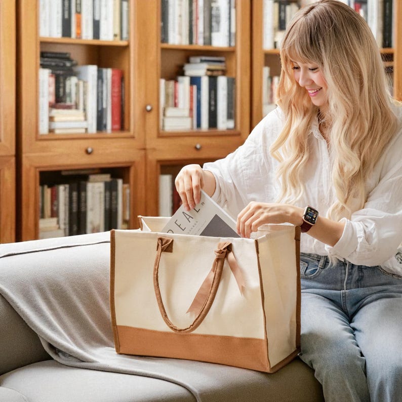 A woman with long blonde hair is sitting on a couch, holding a book and looking inside a large tote bag with a brown handle.