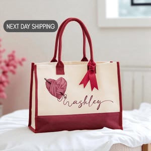 May include: Cream-colored tote bag with burgundy accents and handles. The bag features a heart-shaped yarn design with a knitting needle and the name "Ashley" in cursive. The bag is made of canvas-like material.