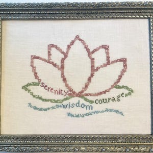 May include: A framed cross-stitch embroidery on off-white fabric featuring a pink lotus flower outline. The words serenity, wisdom, and courage are stitched in green and blue thread below the flower. The piece is enclosed in a silver frame.