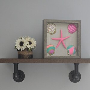 May include: A decorative shelf with a rustic wooden finish, supported by black pipe fittings. It features a framed display of seashells and a pink starfish, alongside a pot of sunflowers.