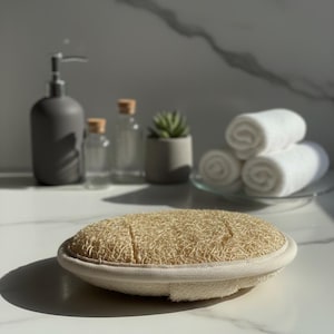 Può includere: Una spugna di luffa naturale con un bordo color crema è appoggiata su una superficie bianca. La luffa è marrone chiaro e ha una superficie strutturata. Altri accessori per il bagno sono sullo sfondo, tra cui un dosatore di sapone grigio e asciugamani bianchi arrotolati.