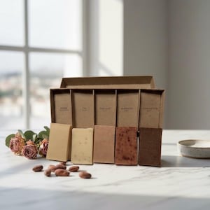 以下が含まれることがあります： 「Aidan」「Fig」「Fine Clay」「Aloe Vera」「Cocoa Butter」と書かれた長方形の石鹸が6つ入った段ボール箱。石鹸はベージュや茶色の様々な色合い。箱の近くにはドライローズとカカオ豆が置かれている。