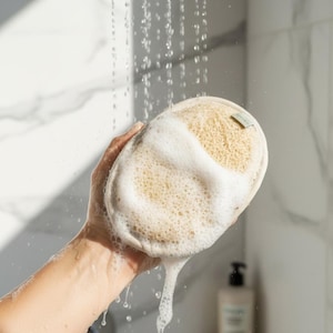Pode incluir: Uma esponja de bucha natural coberta de espuma branca, segurada sob água corrente. A esponja bege é redonda e texturizada, com uma pequena etiqueta anexada. Gotas de água escorrem, criando um efeito espumoso. Uma garrafa de sabão é visível ao fundo.