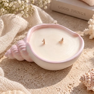 Peut inclure: Un bougeoir en forme de coquillage avec un extérieur rose tendre et un intérieur blanc lisse. La bougie présente deux mèches centrées dans la cire blanche. Le coquillage possède un design en spirale sur un côté et un large bassin ouvert.