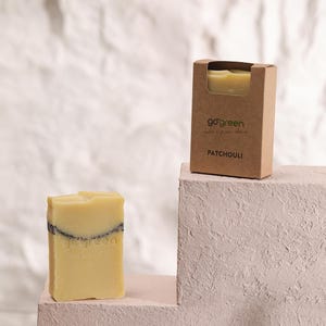 Peut inclure: Deux savons au patchouli, l'un dans une boîte en carton et l'autre présenté séparément. Le savon est jaune clair avec une bande foncée. La boîte porte les mots "go green" et "patchouli".