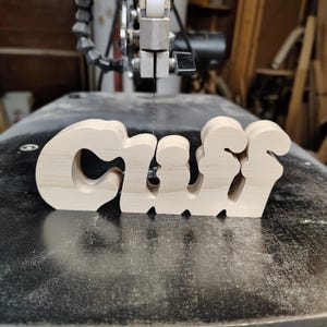 May include: A wooden sign spelling out the name "Graff" in a stylized font. The letters are cut from light-colored wood and stand upright on a dark surface. The sign is in front of a band saw.