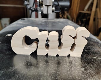 May include: A wooden sign spelling out the name "Graff" in a stylized font. The letters are cut from light-colored wood and stand upright on a dark surface. The sign is in front of a band saw.