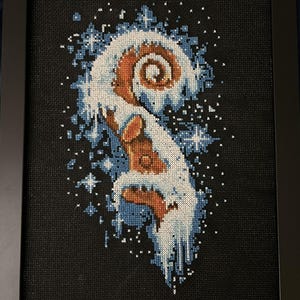 May include: Framed cross-stitch art featuring a stylized design in shades of brown, white, and blue. The artwork depicts a swirling, organic shape with a snowy or icy effect, set against a black background. The piece is framed in a black frame.