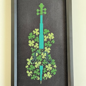 May include: Cross-stitch art of a cello with a shamrock design. The cello is teal, and the shamrocks are various shades of green. The artwork is framed in a black rectangular frame, set against a dark gray background.
