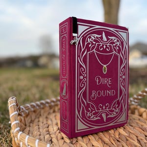 May include: A burgundy book titled "Dire Bound" with silver foil lettering and decorative accents. The book has a black velvet bookmark with a silver charm. The book is displayed on a woven basket.