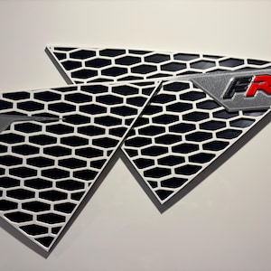 May include: Two triangular car emblems with a black honeycomb pattern and white borders. Each emblem features a silver section with the letters "FR" in red. The emblems are set against a white background.