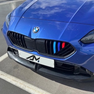 Può includere: Primo piano della parte anteriore di un'auto BMW blu. La griglia nera presenta strisce verticali rosse, blu e azzurre. Il logo BMW è visibile. Una targa bianca con le lettere "AN+" è sotto.