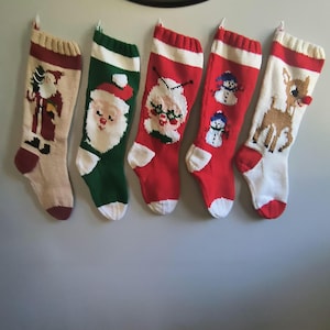 May include: Five knitted Christmas stockings hanging in a row. The first is beige with a Santa figure, the second is green with a Santa face, the third is red with a Mrs. Claus face, the fourth is red with two snowmen, and the fifth is white with a reindeer.