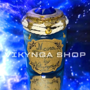 May include: A blue glass bottle with a decorative top and a blue gemstone. The bottle has a gold-colored rim with dripping accents and a label that reads "Mena by Vikynga". The background is a blurred blue and white.