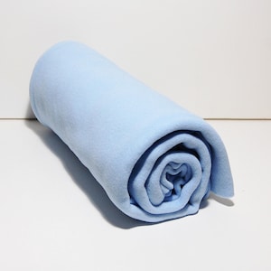 May include: A rolled-up, light blue fleece blanket. The blanket is tightly rolled, revealing the texture of the soft fabric. The color is a pale, sky blue, and the blanket appears to be new and unused. The background is a plain white surface.