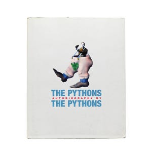 May include: A white book cover with a cartoon illustration of a man standing inside a pair of oversized pants. The text "THE PYTHONS AUTOBIOGRAPHY BY THE PYTHONS" is printed in blue and red letters.