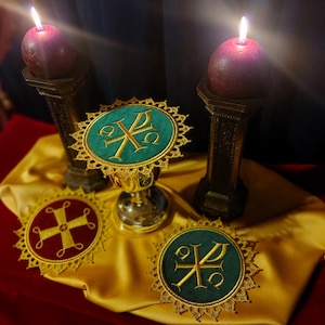 May include: A religious display featuring two lit purple candles on ornate stands. A chalice and three embroidered emblems with gold detailing and teal and red backgrounds are arranged on a gold cloth.