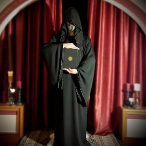 May include: A person in a black hooded robe holding a book with a gold emblem. The robe has decorative trim and is worn in front of a red curtain. The scene is lit by candles on small wooden tables.