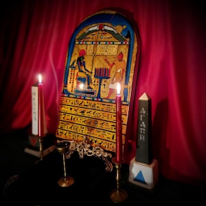 May include: An Egyptian-themed display. A large, ornate, painted tablet with hieroglyphs and figures is flanked by lit red candles in brass holders. A small black obelisk with white text and a gold incense burner complete the scene.