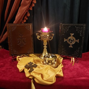 Könnte beinhalten: Eine verzierte religiöse Darstellung mit einer brennenden Kerze in einem goldfarbenen Halter, flankiert von zwei ledergebundenen Büchern mit geprägten Kreuzen. Ein kleines Kreuz und eine dekorative Box liegen auf einem drapierten Tuch.