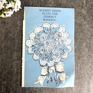 May include: A vintage book cover with the title "MODERN BRIDE PLANS THE PERFECT WEDDING" in blue text. The cover features a detailed illustration of a bouquet of white and light blue flowers tied with a white ribbon. The background is light blue.