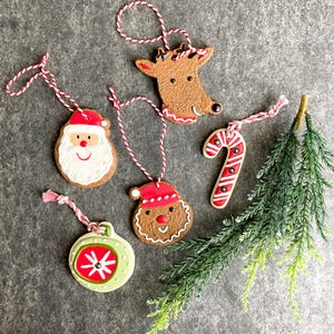 May include: Five Christmas ornaments made of gingerbread with red and white icing. The ornaments are shaped like a reindeer, Santa Claus, a candy cane, a snowman, and a Christmas tree ornament. The ornaments are hanging from red and white twine.