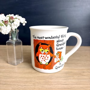 May include: White ceramic mug with a handle, featuring a cartoon owl design. The mug has the text "the most wonderful thing about Grandpa's house... is Grandpa!" in black and orange. A small glass vase with white flowers is to the left.