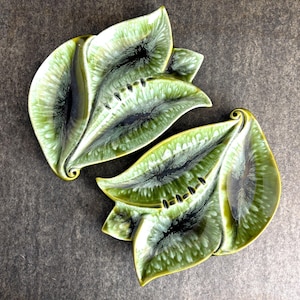 May include: Two leaf-shaped ceramic dishes with a glossy, green glaze. The dishes have a dark, almost black, center and a textured surface. The edges are a lighter green, and the overall design is organic and decorative.