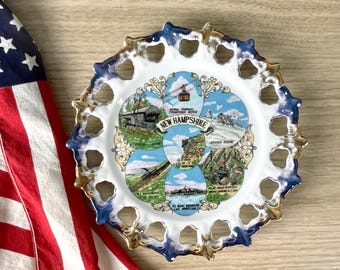 May include: A decorative plate featuring scenes from New Hampshire, with a blue and gold rim. The plate has a white center with images of the state's landmarks and the text "NEW HAMPSHIRE". An American flag is partially visible in the background.
