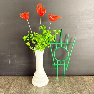 May include: A white vase holds green foliage and red flower and bird decorations. A green metal trellis stands beside the vase. The items are set against a dark background.