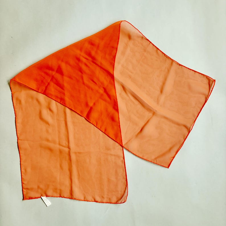 May include: A two-tone scarf in shades of orange. The scarf has a rectangular shape with a diagonal colour block design. The top section is a vibrant orange, while the bottom section is a lighter peach colour. The edges are finished with a thin, darker orange trim.