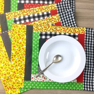 May include: A white ceramic plate and a silver spoon rest on a colorful patchwork placemat. The placemat features various fabric patterns, including floral, polka dot, and gingham, in red, yellow, green, and black.